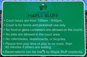 Green sign on a fence with tennis and pickleball court rules for Maple Bluff, listing hours, prohibited items, and reservation info. White text and a maple leaf logo appear at the top.