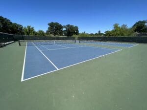 An empty outdoor tennis court with a blue playing surface and green surrounding area, surrounded by a black fence, under a clear blue sky with trees in the background.