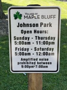 Sign for Johnson Park in Maple Bluff showing open hours: Sunday-Thursday 5am-11pm, Friday-Saturday 5am-12am. Amplified noise prohibited between 9pm-7am. Sign is posted on grass with some trees in background.