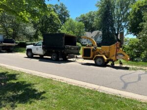 A white utility truck with a dark cargo bed is parked on a residential street, hitched to a yellow wood chipper. Trees and houses are visible in the background on a sunny day.