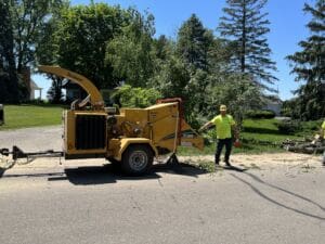 A worker in a yellow shirt and hard hat stands next to a yellow wood chipper on a sunny day, with branches and leaves on the ground and trees in the background.