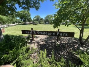 A wooden sign reading Neal H. Stoddard Park stands at the edge of a grassy park, surrounded by green trees and shrubs on a sunny day.
