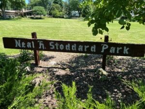 A wooden sign reading Neal K. Stoddard Park stands in front of a grassy field with trees and houses in the background on a sunny day.