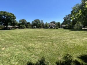 A large, open grassy field bordered by trees and houses under a clear blue sky on a sunny day. Shadows from nearby trees are visible in the foreground.