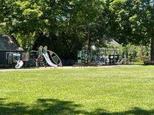 A sunny park with green grass, large trees, and a playground featuring slides, climbing structures, and swings. A few benches are nearby, and a small building is visible on the left.