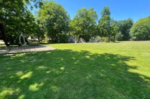 A sunny park with green grass, large trees, and a playground area with swings and slides. Shadows of trees cover parts of the lawn, and a building is visible in the background.