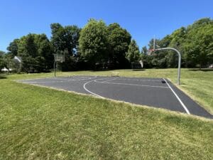 An outdoor basketball court with two hoops sits on a grassy field surrounded by trees under a clear blue sky. There are no people present, and the scene is bright and sunny.