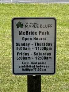 A sign in McBride Park, Village of Maple Bluff, shows open hours: Sunday-Thursday 5:00am–11:00pm, Friday-Saturday 5:00am–12:00am. Amplified noise is prohibited 9:00pm–7:00am.