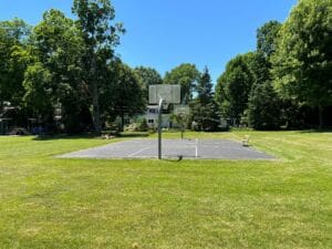 A sunny outdoor basketball court surrounded by green grass and tall trees, with houses in the background. The court has two basketball hoops, and a bench sits nearby.