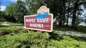 A wooden sign reading “Maple Bluff Marina” with an illustration of a boat stands among green shrubs and trees, with a paved path and water visible in the background on a sunny day.