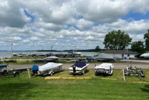 Several small boats, some covered, sit on grassy and gravel areas near a lake with a dock. The sky is partly cloudy, and trees border the water in the background.