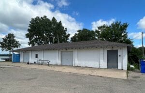 A long, white, cinder block building with a gray roof sits on a cracked concrete lot. There are metal racks, a blue portable toilet, and trees in the background under a partly cloudy sky.
