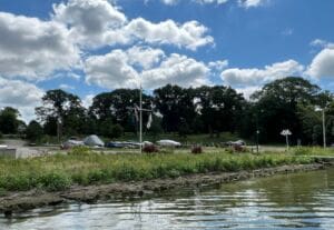 A small marina with boats, cars, and tents along a grassy shoreline. Trees line the background under a blue sky with scattered clouds, and the water in the foreground is calm.