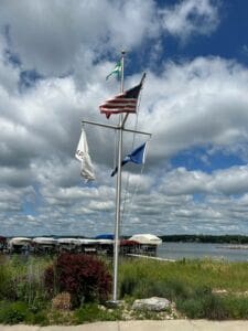A flagpole with four flags, including the American flag, stands near a waterfront with docked boats, green grass, and shrubs under a partly cloudy sky.