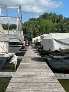 A wooden dock walkway leads between rows of covered boats at a marina, surrounded by green trees under a partly cloudy sky.