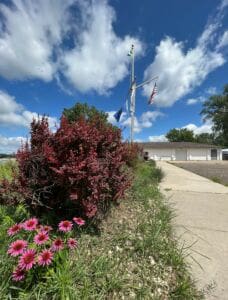 Pink coneflowers and red shrubs grow beside a sidewalk leading to a building with three flags, including the American flag, flying on flagpoles under a bright blue sky with scattered clouds.