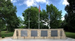A stone memorial wall with plaques and military insignias stands before three flagpoles, the center one flying the American flag. Lush green trees and a blue sky with scattered clouds fill the background.