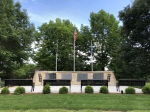 A memorial with stone plaques, three flagpoles (one with a U.S. flag), black benches, and circular emblems. Lush green trees and grass surround the area under a partly cloudy sky.