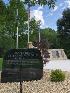 A stone marker reading Maple Bluff Veterans Memorial stands in front of a memorial wall, with three flagpoles and trees in the background under a partly cloudy sky. The marker lists honorees and dedication details.
