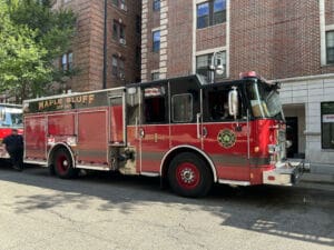 A red fire truck labeled Maple Bluff Engine 1 is parked on a city street beside brick buildings, with sunlight casting shadows on the vehicle and the pavement.