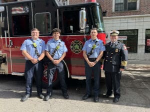 Four firefighters, three in light blue shirts and dark pants, and one in a black dress uniform with a hat, stand smiling in front of a red fire truck parked on a street.