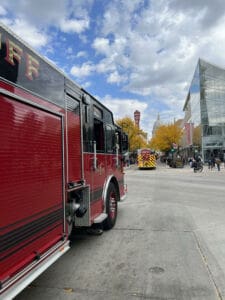 A large red fire truck is parked on a city street with another emergency vehicle ahead. Pedestrians walk nearby, and buildings with glass windows and autumn trees line the street under a partly cloudy sky.