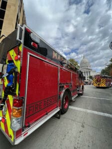 A red fire rescue truck labeled “Maple Bluff” is parked on a city street near another fire truck, with the white dome of a government building visible under a partly cloudy sky.