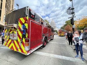 A red fire truck labeled MAPLE BLUFF drives down a city street during a parade. People stand on the sidewalk watching, and the Wisconsin State Capitol is visible in the background under a partly cloudy sky.