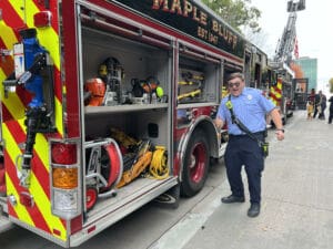 A firefighter in uniform smiles and gives a thumbs up beside an open fire truck compartment filled with equipment, including hoses, tools, and safety gear, on a city street.
