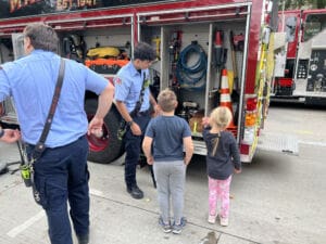 Two firefighters show equipment stored in a fire truck to a young boy and girl. The children stand near the open truck compartments, looking at tools and hoses. The scene takes place outdoors on a paved area.