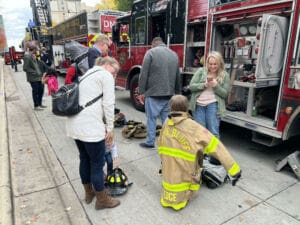 Adults and children stand next to a fire truck parked on the street. A child wears a large firefighters jacket and helmet while an adult kneels nearby. Other people interact with firefighters and equipment.