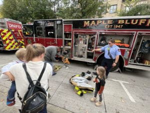 A fire truck is parked on the street with its side compartments open. Firefighters interact with children and adults nearby; one firefighter points while another kneels, and a child stands among firefighting gear on the ground.