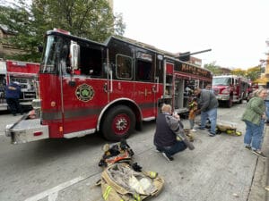 A red fire truck labeled Maple Bluff is parked on a street with its side compartments open. Several people, some in firefighting gear, are gathered around the truck, with equipment and clothing on the ground nearby.