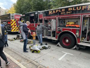 People explore a Maple Bluff fire truck parked on a street. Firefighting gear is laid out on the ground, and a child interacts with two adults near the truck. Other people are visible in the background. Trees line the street.