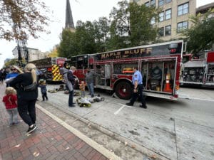 A Maple Bluff fire truck is parked on a city street during an event. Firefighters stand nearby, while families with children observe and interact. Firefighting gear and equipment are displayed on the ground. Trees and buildings are in the background.