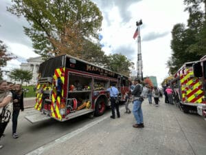 A Maple Bluff fire truck is parked on a street during a community event. People, including families and firefighters, gather around. Trees and buildings are in the background, and an American flag is raised on a tall ladder.
