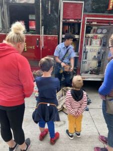 A firefighter shows equipment from a fire truck to a group of children and adults standing on a sidewalk, engaging them in a demonstration.