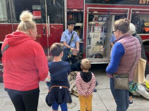A firefighter in uniform talks to a group of children and adults standing beside an open fire truck, showing equipment inside. The group listens attentively on a sidewalk.