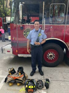 A firefighter stands smiling in front of a red fire truck, wearing a blue uniform. Firefighting gear, including boots, a helmet, jacket, gloves, and a mask, is neatly arranged on the ground in front of him.