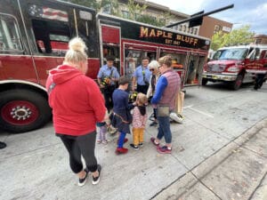 A group of children and adults gather around firefighters next to a fire truck labeled Maple Bluff parked on a city street during an outdoor event. The firefighters show equipment to the kids.