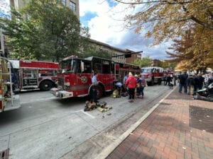 A group of people gathers on a city sidewalk near fire trucks parked on the street. Firefighters are working with gear on the ground as bystanders watch. Autumn trees line the street in the background.