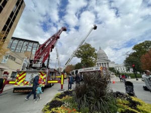 Fire trucks with extended ladders are parked near the Wisconsin State Capitol. Firefighters and civilians, including children, are present. Its a cloudy day with people walking and observing the scene.