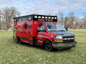 A red Maple Bluff Fire Rescue ambulance is parked on grass, with trees and houses in the background. The vehicle features yellow and green lettering, emergency lights, and a prominent Chevy logo on the front grille.