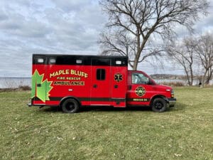 A red Maple Bluff Fire Rescue ambulance is parked on grass near leafless trees and a body of water under a cloudy sky. The vehicle features a green maple leaf graphic and the words “Rescue 56.”.