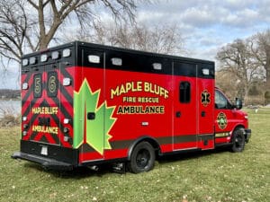 A red and black ambulance labeled Maple Bluff Fire Rescue Ambulance is parked on grass near leafless trees and a body of water, with a large green maple leaf graphic on the side.