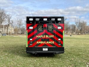 The rear view of a Maple Bluff ambulance numbered 56, parked on grass with red and green chevron stripes, surrounded by leafless trees and houses under a partly cloudy sky.
