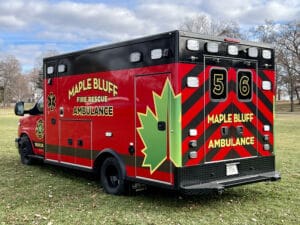 A red and black Maple Bluff Fire Rescue ambulance, marked with a large green maple leaf and the number 56, is parked on grass with trees and cloudy sky in the background.