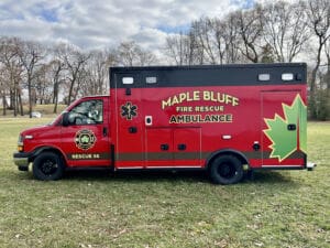 A red Maple Bluff Fire Rescue ambulance with a large green maple leaf design is parked on grass in a park, with trees and a cloudy sky in the background.