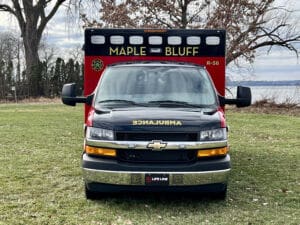 A red and black ambulance labeled Maple Bluff is parked on grass near trees and a lake, facing directly toward the camera.