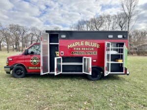 A red Maple Bluff Fire Rescue ambulance parked on grass with its side doors open, revealing storage compartments and equipment, against a backdrop of leafless trees and a partly cloudy sky.